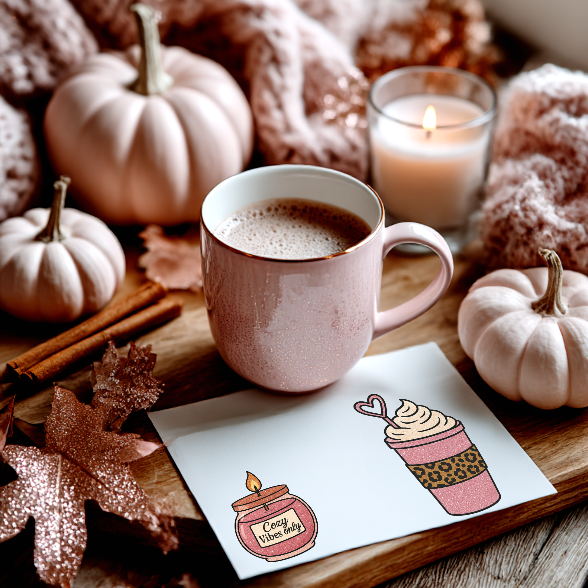 Pink mug of hot chocolate with pumpkins, cinnamon, and a lit candle on a wooden surface.