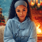 Woman wearing a light blue hoodie with 'Your Design Here' text, sitting in front of a Christmas tree and fireplace.