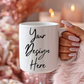 White mug with 'Your Design Here' text held by a hand with glittery nail polish against a festive background.